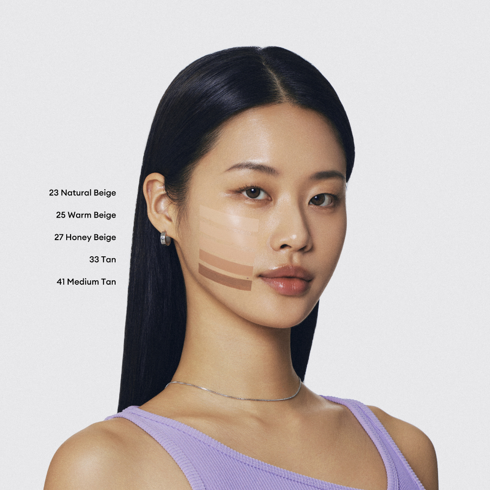 A young woman with warm undertones and dark hair looks at the camera. Swatches of five different foundation shades are visible on her cheek. A young woman with warm undertones and dark hair looks at the camera. Swatches of five different foundation shades are visible on her cheek.