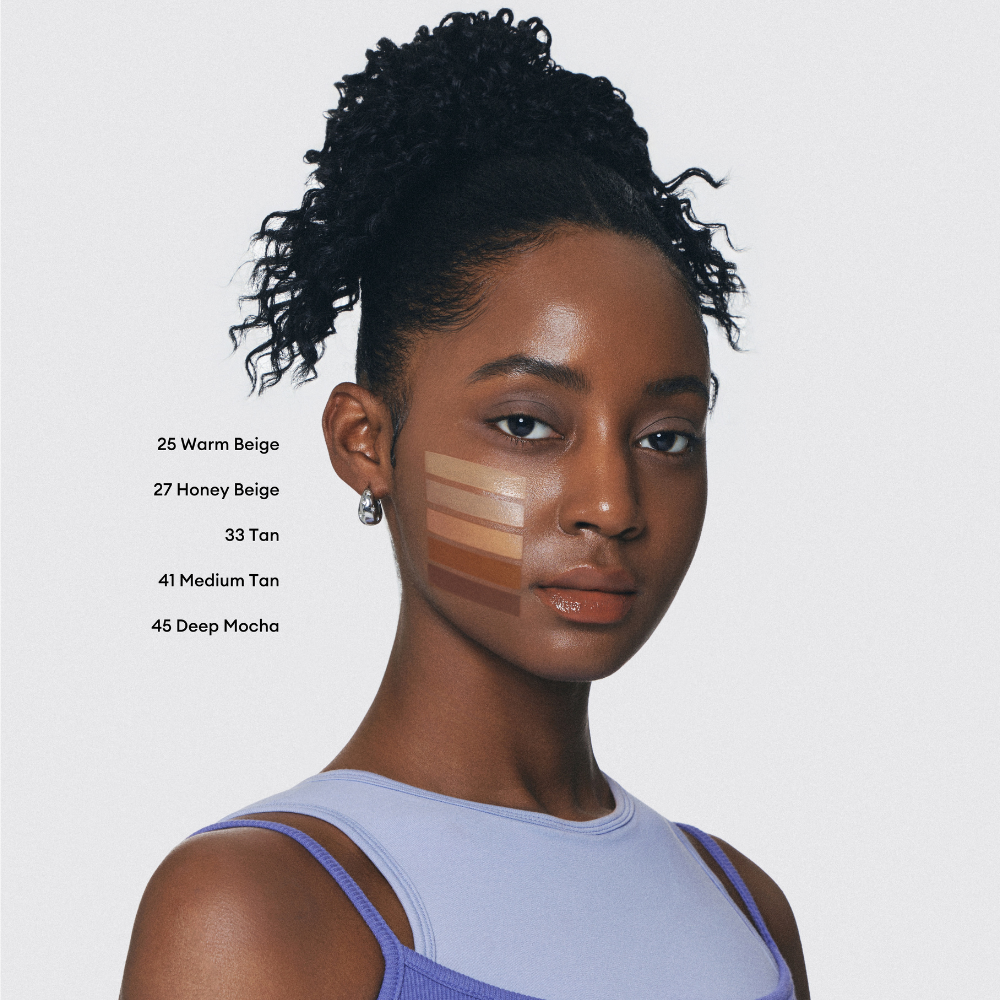A young woman with dark skin and curly hair looks at the camera. Swatches of five different foundation shades are visible on her cheek. A young woman with dark skin and curly hair looks at the camera. Swatches of five different foundation shades are visible on her cheek.