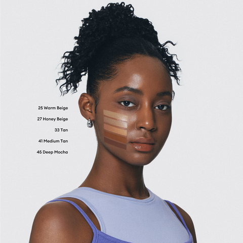 A young woman with dark skin and curly hair looks at the camera. Swatches of five different foundation shades are visible on her cheek. A young woman with dark skin and curly hair looks at the camera. Swatches of five different foundation shades are visible on her cheek.