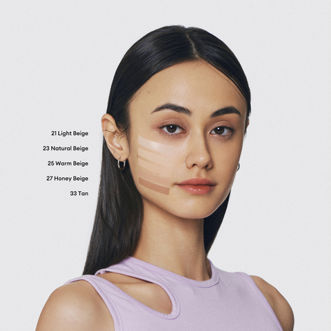 A young woman with medium brown skin and dark hair looks at the camera. Swatches of five different foundation shades are visible on her cheek. A young woman with medium brown skin and dark hair looks at the camera. Swatches of five different foundation shades are visible on her cheek.