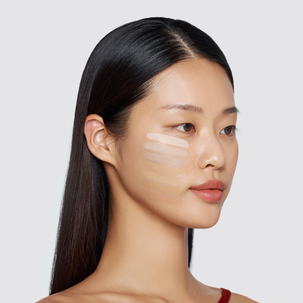 A woman with long, dark hair has a series of five different foundation shades swatched in horizontal stripes on her cheek. A woman with long, dark hair has a series of five different foundation shades swatched in horizontal stripes on her cheek.