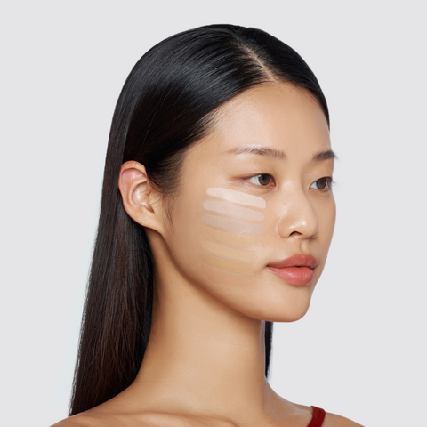 A woman with long, dark hair has a series of five different foundation shades swatched in horizontal stripes on her cheek. A woman with long, dark hair has a series of five different foundation shades swatched in horizontal stripes on her cheek.