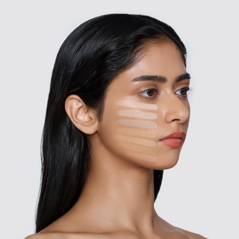 A woman with long, dark hair has a series of five different foundation shades swatched in horizontal stripes on her cheek. A woman with long, dark hair has a series of five different foundation shades swatched in horizontal stripes on her cheek.