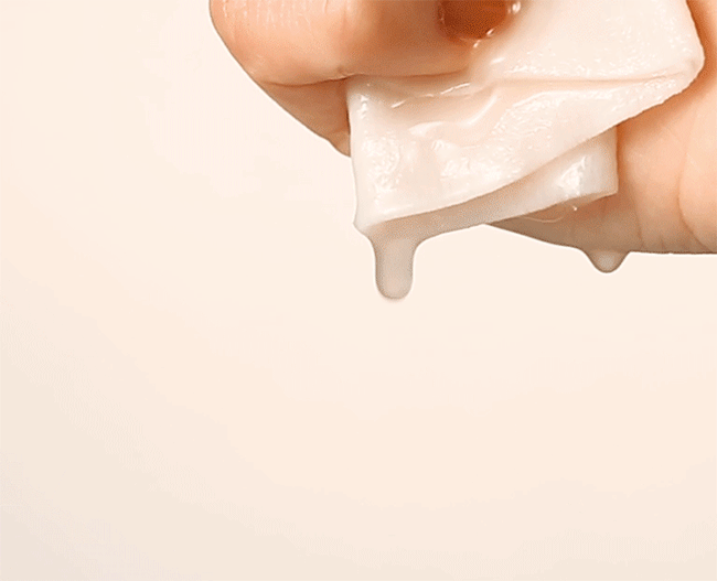 Close-up macro shot of a hand squeezing a white sheet mask material, showing a thick, milky, white serum dripping down. This image highlights the rich, concentrated ampoule serum texture of the Time Revolution Night Repair Ampoule Sheet Mask used for intensive K-beauty skin treatment.