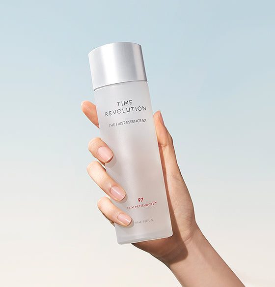 A hand holding a sleek, silver-capped bottle of Time Revolution The First Essence RX against a pastel sky background.