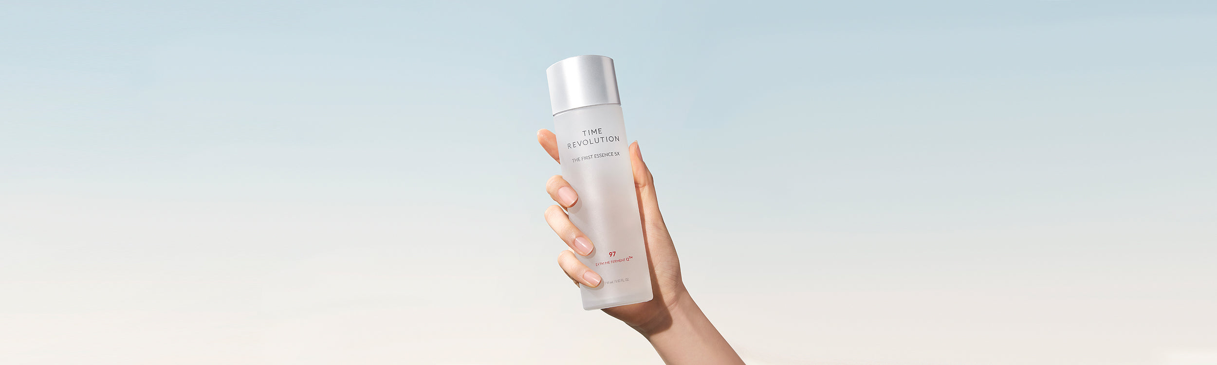 A hand holding a sleek, silver-capped bottle of Time Revolution The First Essence RX against a pastel sky background.