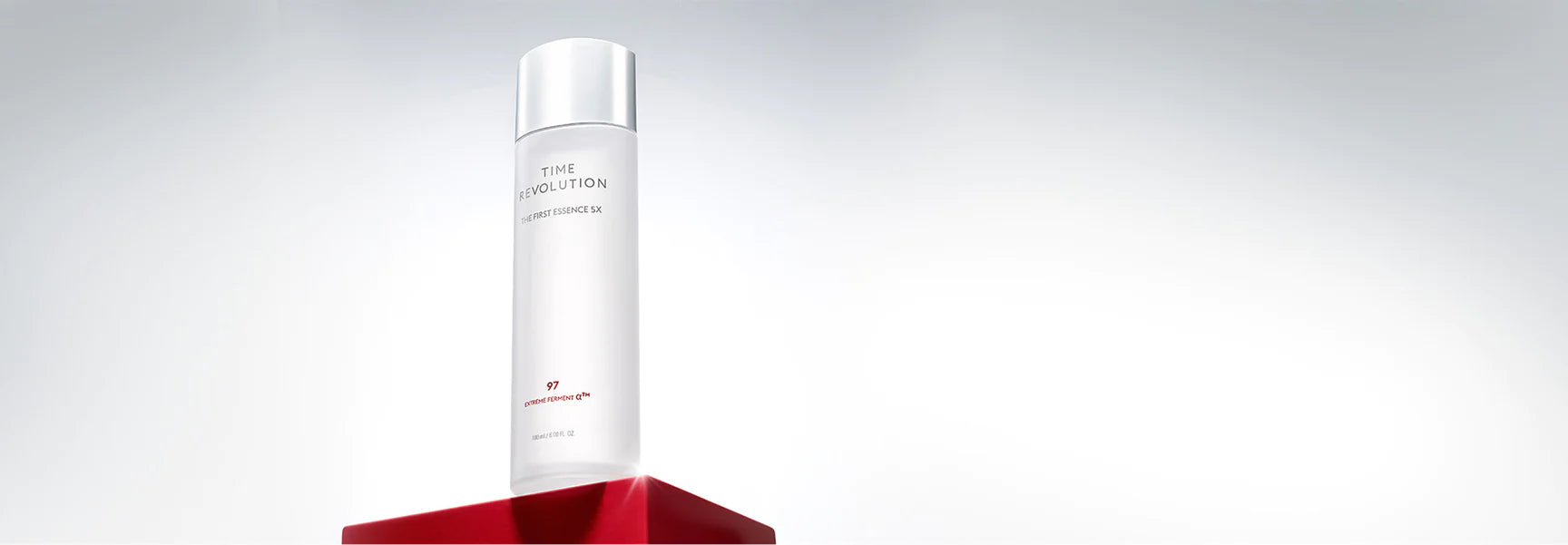 Sleek white Time Revolution The First Essence SX bottle displayed on a bold red pedestal, highlighting premium skincare.