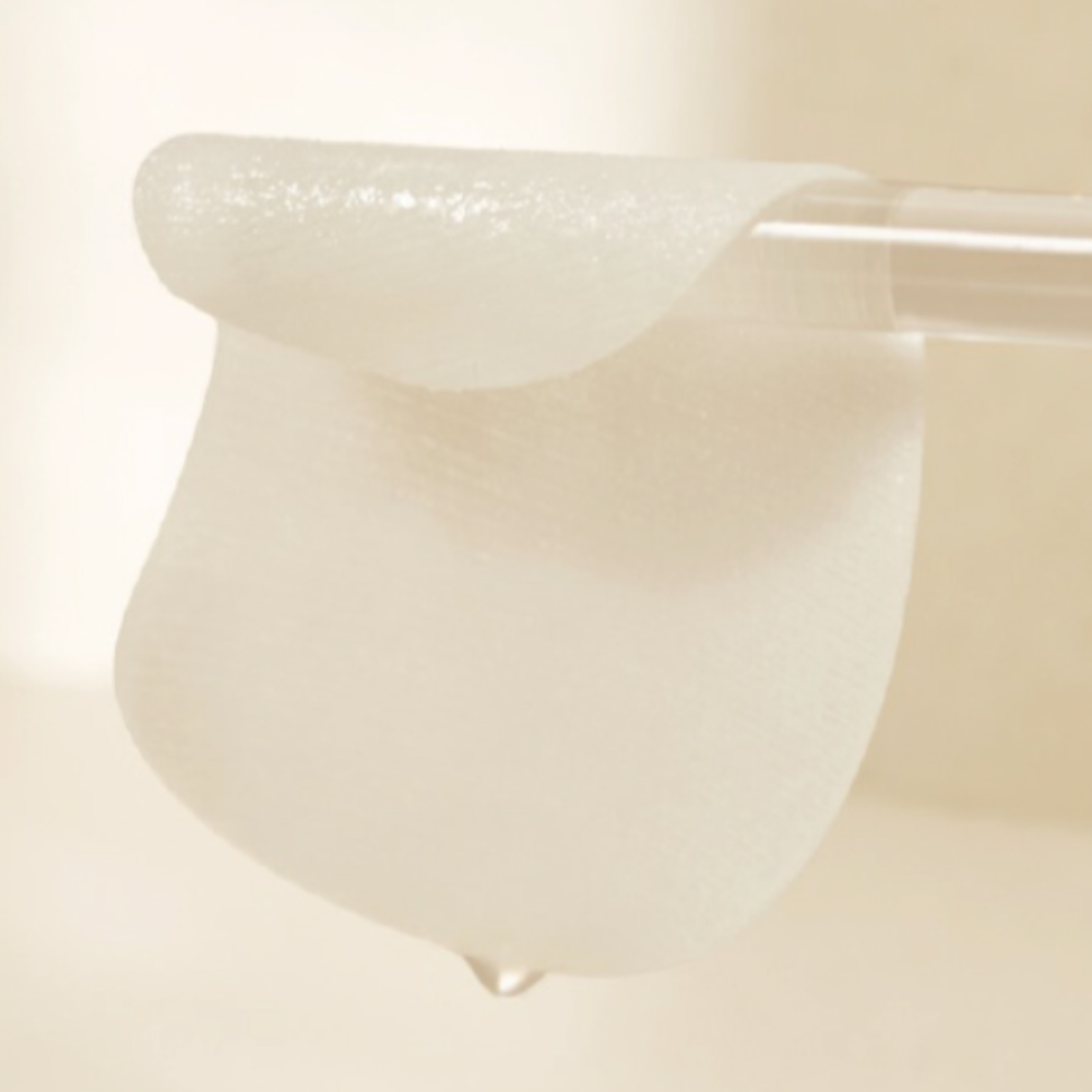 A close-up of a moist skincare pad draped over a clear glass rod, with a droplet of essence falling from the pad, highlighting its hydration and texture.
