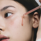 Close-up of a woman applying a pink serum drop from a glass dropper onto their cheek.