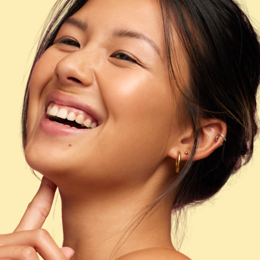 A woman smiling radiantly with glowing, healthy skin and minimal makeup, showcasing a natural dewy complexion against a soft yellow background.