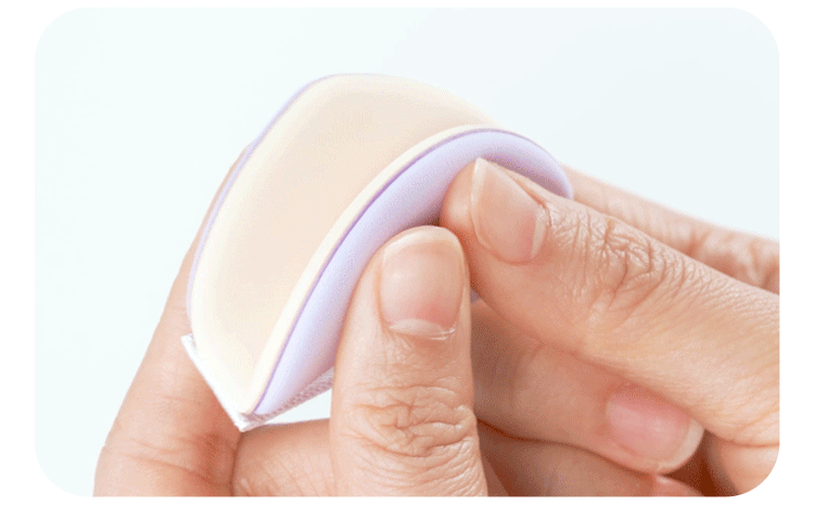 Close-up shot of a hand gently squeezing the soft, thick material of the tear-drop shaped Missha Layering Puff. The puff is beige on one side and light purple on the other, demonstrating its dual-layer structure and flexibility for applying cushion foundation.