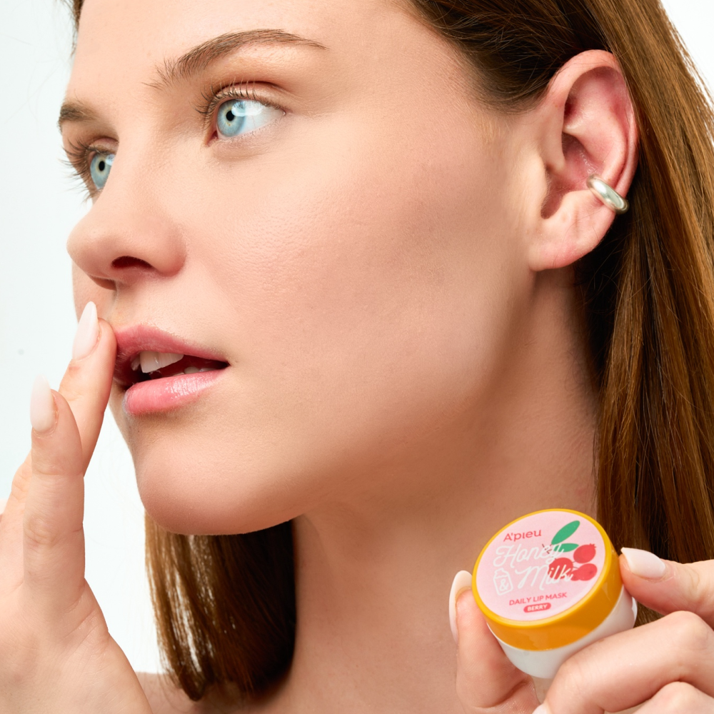Close-up of a model with blue eyes applying A'pieu Honey Milk Daily Lip Mask in Berry flavor with her finger to her lips. The small, yellow and white jar, with a pink label featuring raspberries, is visible in her hand, highlighting the product's use for moisturizing and caring for dry lips.