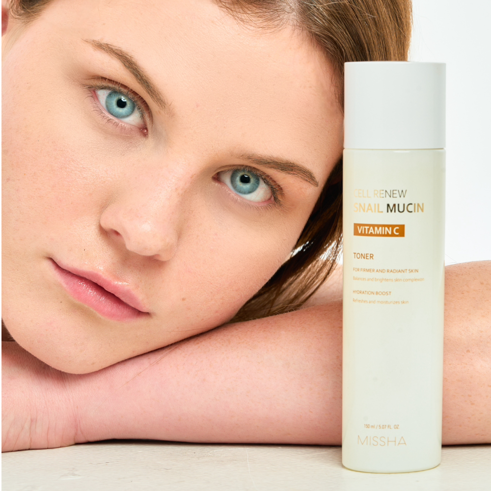 Portrait of a woman with blue eyes resting her head near the full-size bottle of Missha Cell Renew Snail Mucin Vitamin C Toner. The image highlights the brightening benefits of the toner for radiant skin.