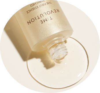 A hand holding a sleek, golden bottle of Time Revolution The First Essence  against a pastel yellow background.