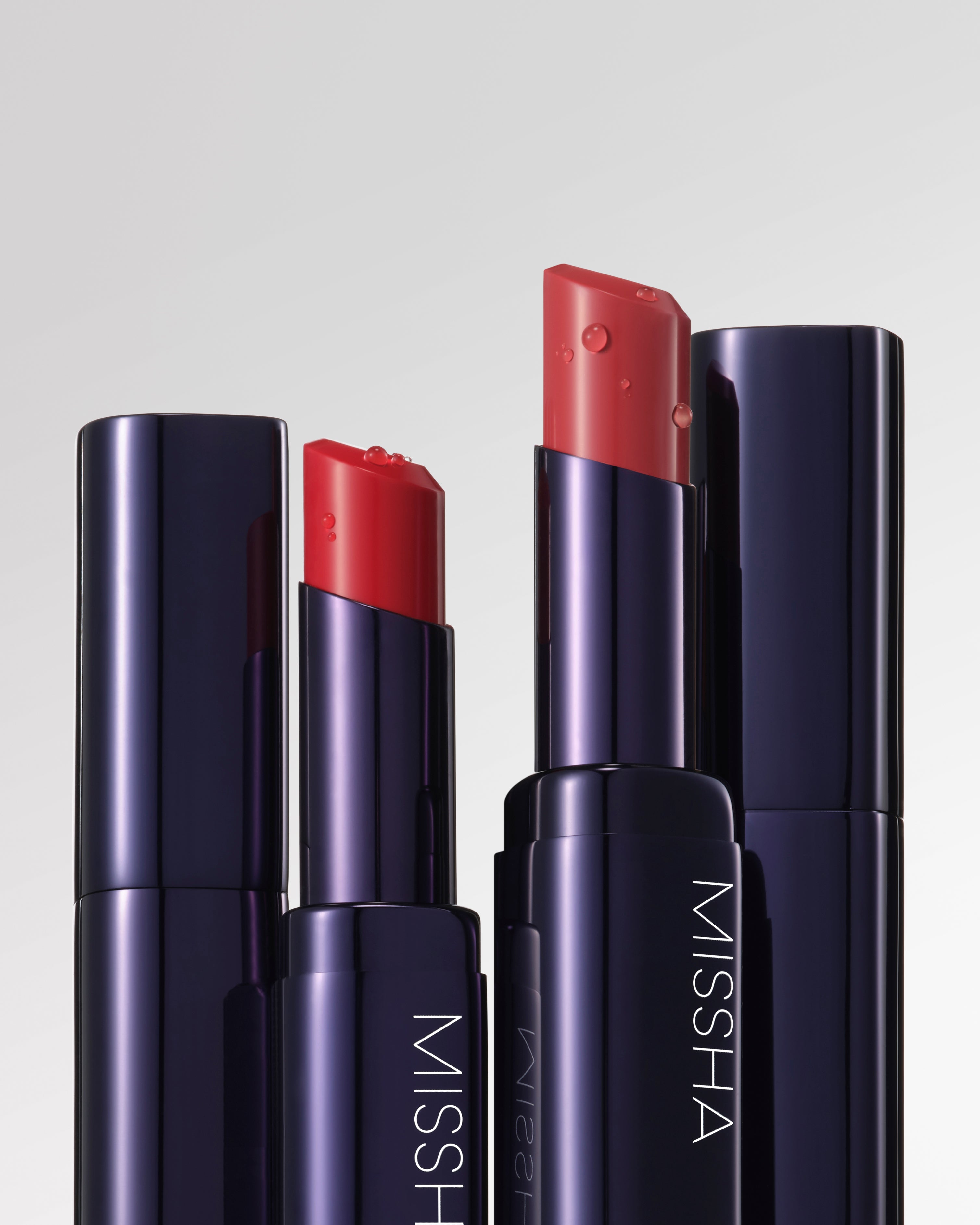 Close-up of sleek MISSHA lipsticks in deep navy cases, featuring rich coral-red shades with a glossy, hydrating texture and visible water droplets emphasizing moisture and shine.