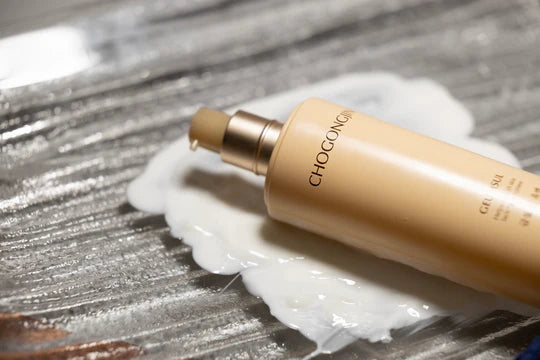A golden skincare bottle lies on a reflective surface with a creamy, white product oozing out beside it.