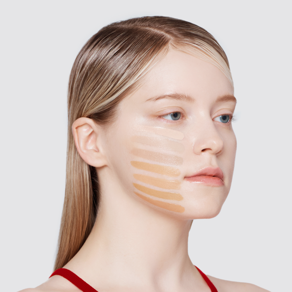 A woman with light blonde hair has a series of six different foundation shades swatched in horizontal stripes on her cheek.