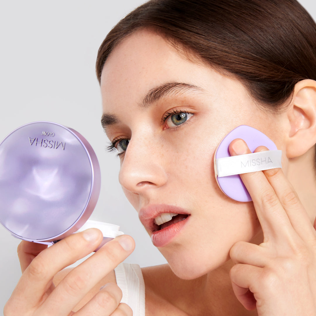 Person applying MISSHA Glow Cushion foundation with a lavender puff while holding a matching compact case, creating a dewy, radiant finish.