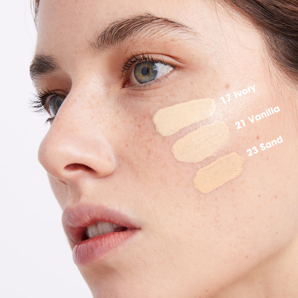 Close-up of a person’s face showing three foundation swatches labeled 17 Ivory, 21 Vanilla, and 23 Sand on their cheek to compare skin tone shades.