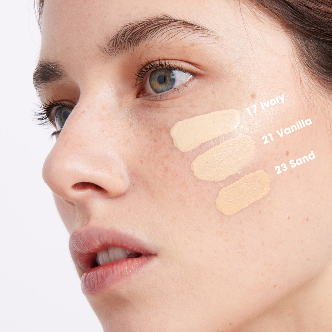 Close-up of a person’s face showing three foundation swatches labeled 17 Ivory, 21 Vanilla, and 23 Sand on their cheek to compare skin tone shades.