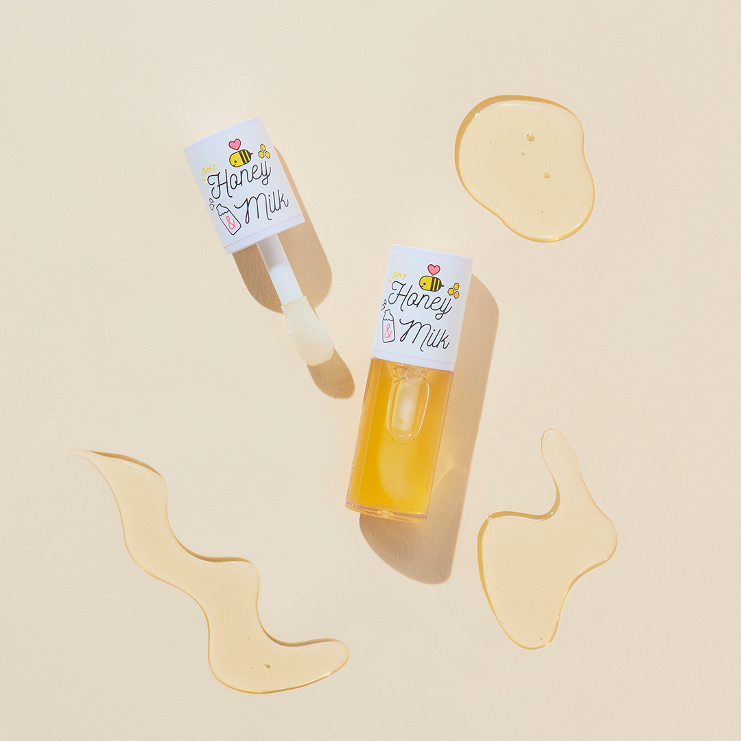 A'pieu Honey & Milk Lip Oil in a clear tube with orange-colored product visible inside. The packaging is adorned with illustrations of honeycomb and the product is equipped with a white applicator.