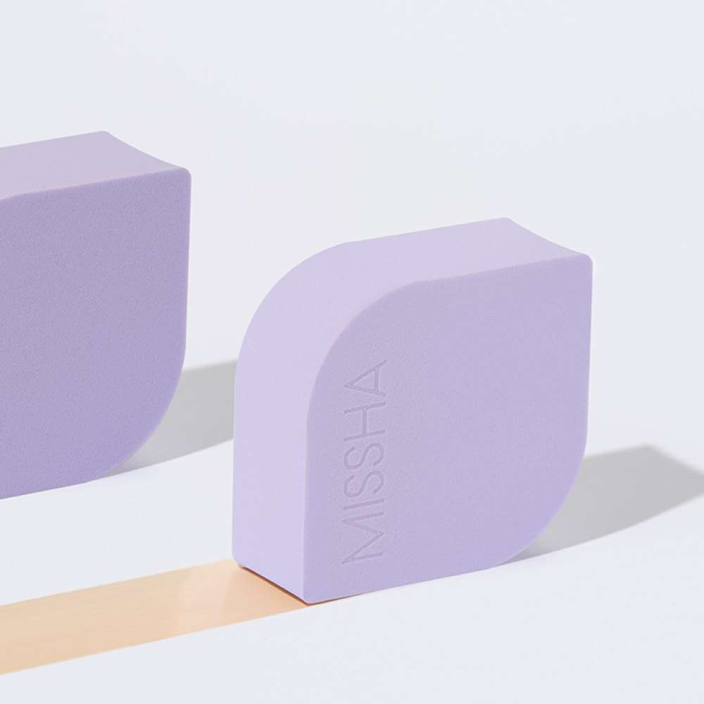 The MISSHA Layering Fit Puff resting on a surface, shown with a smooth, swatched trail of its peach-toned cream formula to demonstrate its blendable texture and coverage.