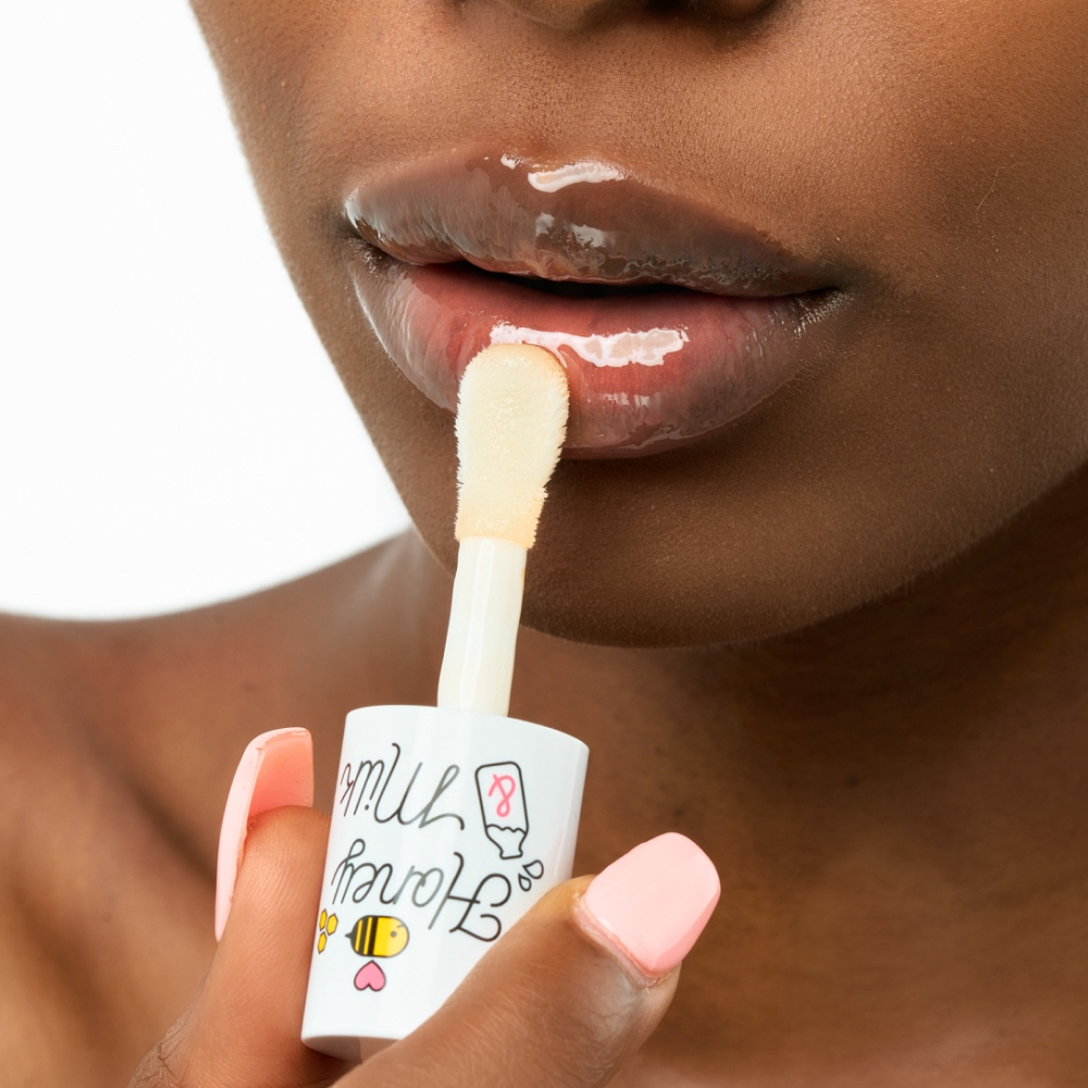 Close-up of a Black model applying the A'pieu Honey & Milk Lip Oil using the oversized doe-foot applicator, which leaves a high-shine, glossy finish on the lips, emphasizing deep moisturizing and plumpness.