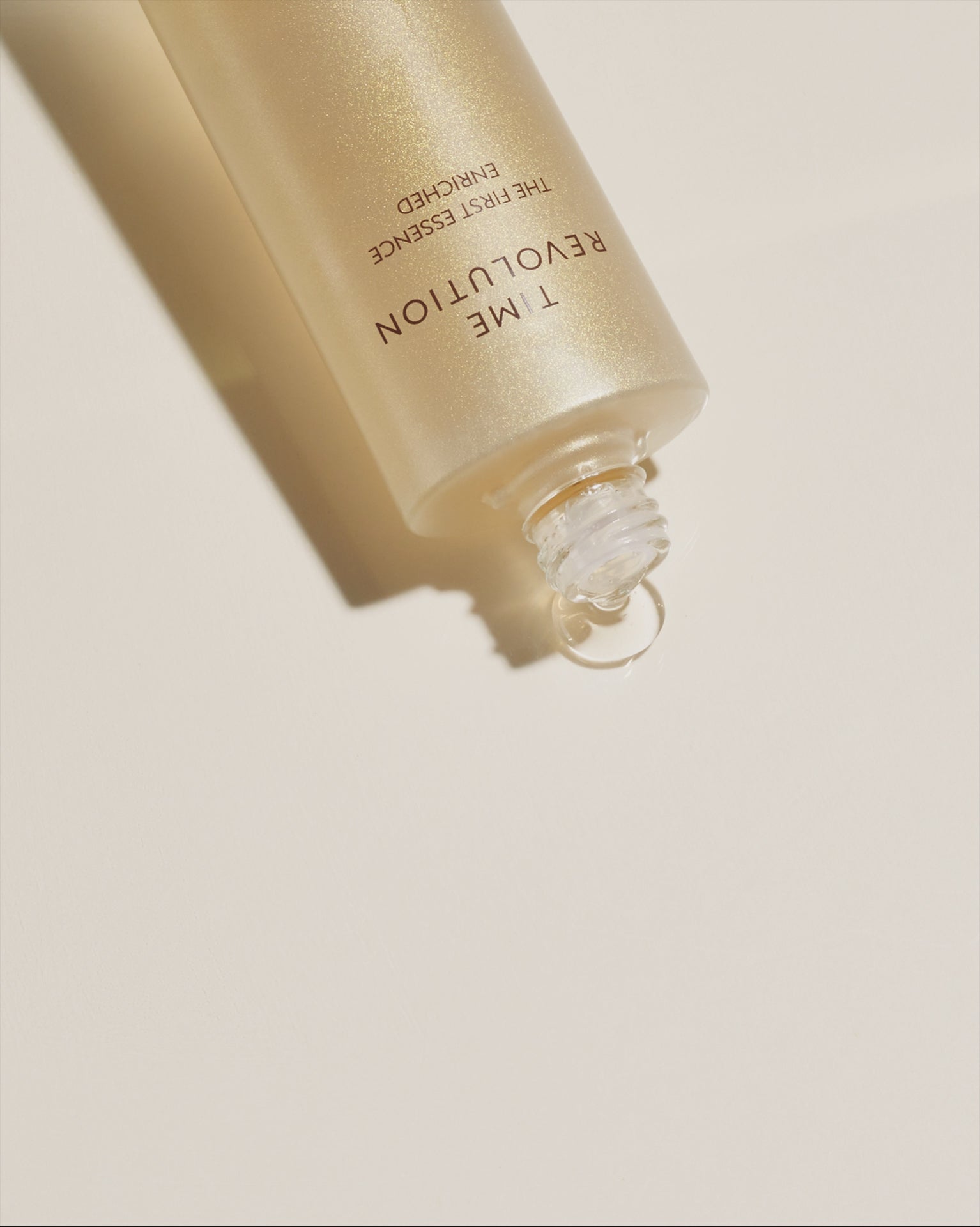 MISSHA Time Revolution The First Essence Enriched bottle lying on a cream surface with liquid essence dripping from the opening, highlighting its luxurious texture and radiant golden glow.