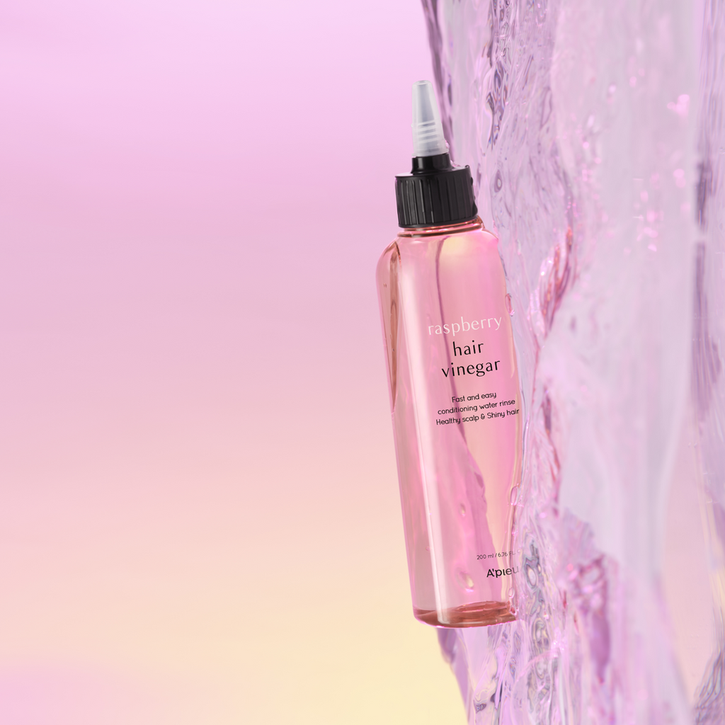 A pink bottle labeled 'hair vinegar' with a black cap and white text, branded as A'pieu. With purplish water flowing next to it along its length. 