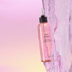 A pink bottle labeled 'hair vinegar' with a black cap and white text, branded as A'pieu. With purplish water flowing next to it along its length. 