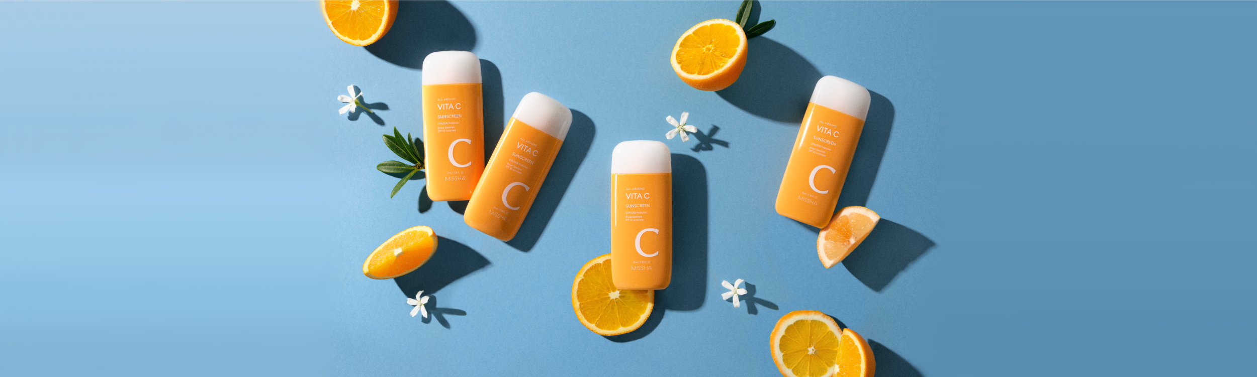Four orange sunscreen bottles are arranged on a blue background, surrounded by fresh orange slices and white flowers.