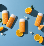 Four orange sunscreen bottles are arranged on a blue background, surrounded by fresh orange slices and white flowers.