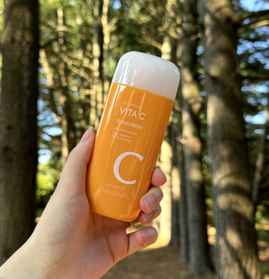 MISSHA VITA C Sunscreen bottle, a daily-use vitamin C-enriched sunscreen with high SPF and PA ratings for strong protection against UVA and UVB rays. The product is held in the hand of a model with the background of a forest.