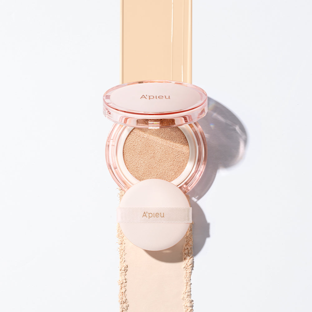 Overhead shot of the open A'pieu Cushion Foundation compact and the accompanying puff, centered on a light beige makeup streak and loose powder, illustrating the full coverage, dewy cream texture.