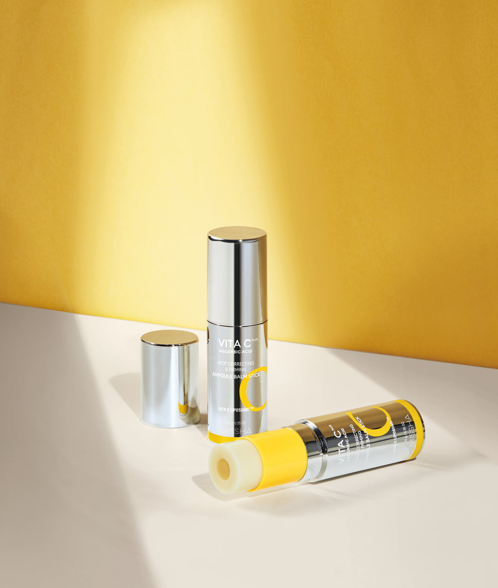Two silver Missha Vita C Plus Spot Correcting & Firming Ampoule Balm Sticks against a sunny yellow background. One stick is upright, and the other is lying down with the yellow Vitamin C balm exposed. This image showcases the portable, roll-on balm stick packaging.