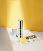 Two silver Missha Vita C Plus Spot Correcting & Firming Ampoule Balm Sticks against a sunny yellow background. One stick is upright, and the other is lying down with the yellow Vitamin C balm exposed. This image showcases the portable, roll-on balm stick packaging.