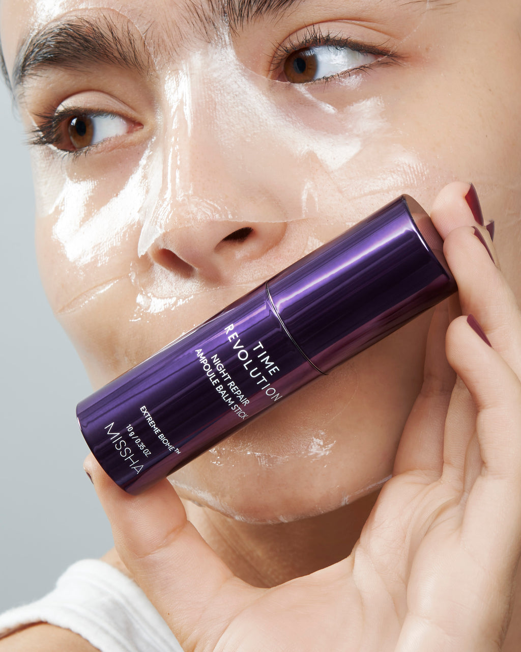 Model Holds Missha Time Revolution Night Repair Ampoule Balm Stick to her cheek and under-eye area. This hydrating, anti-aging stick is infused with Extreme Biome (10 probiotics) and collagen to target fine lines, improve elasticity, and provide a radiant glow on the go. She has the Hydro Gel Mask Applied