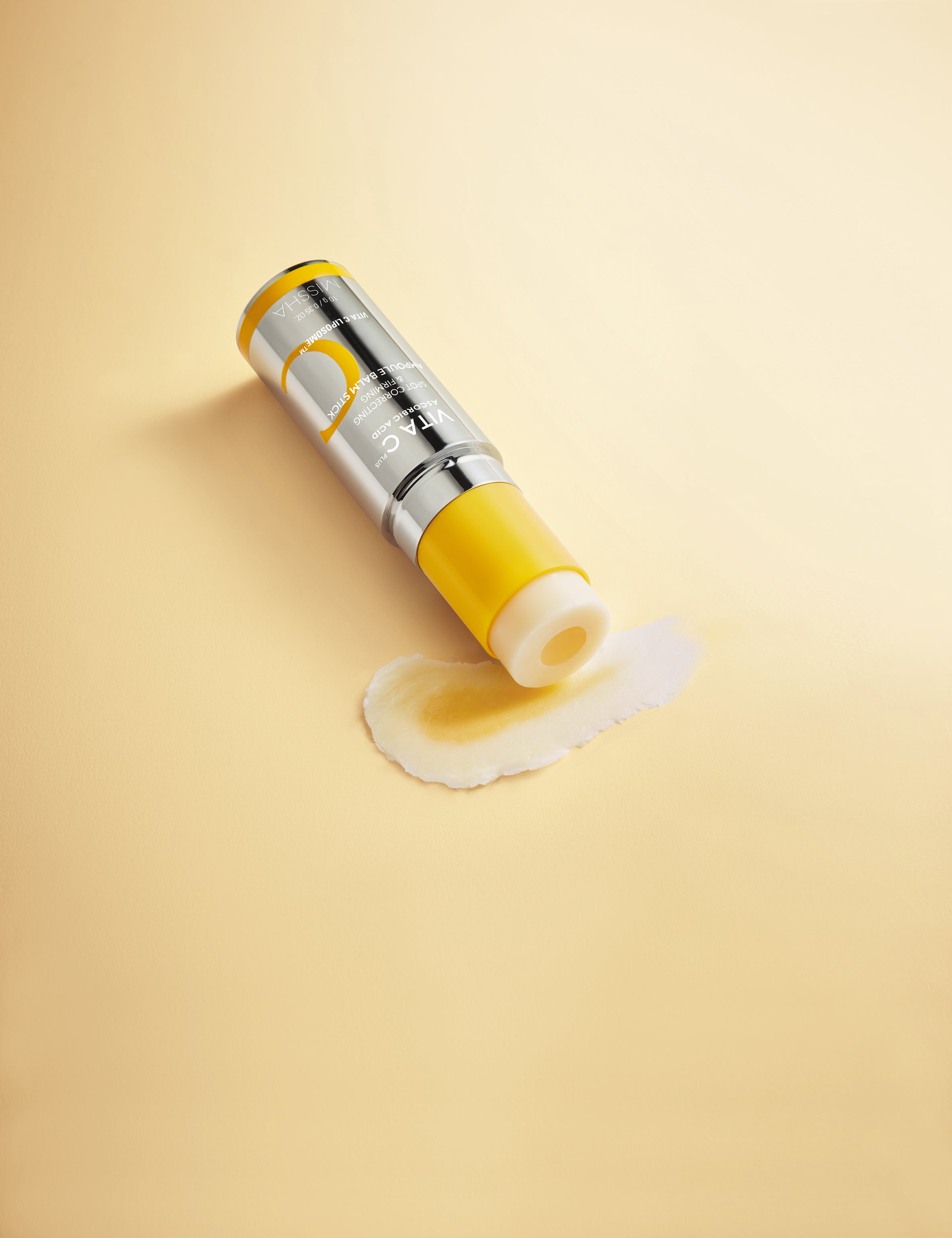 MISSHA Vita C Plus Spot Correcting Ampoule lying on a soft yellow surface with a smear of product, showcasing its brightening vitamin C formula and creamy texture.