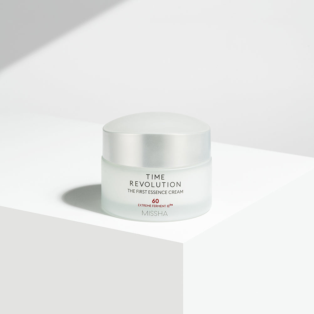 A jar of Missha Time Revolution The First Essence Cream placed on a white surface with a shadow falling across the frame.