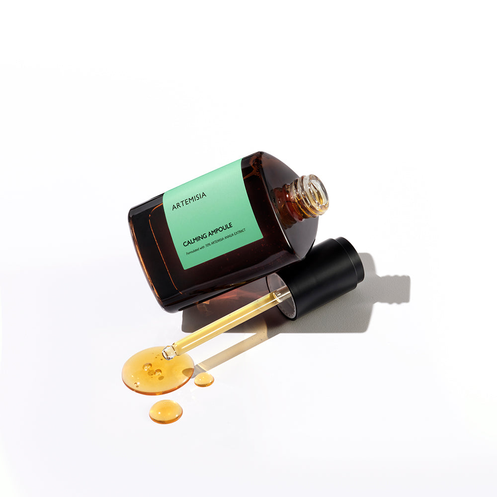 A dark amber bottle of the Missha Artemisia Calming Ampoule lying on its side, with the black-capped dropper resting in a pool of the golden-yellow serum. The image highlights the texture and packaging of the soothing Mugwort skincare product.