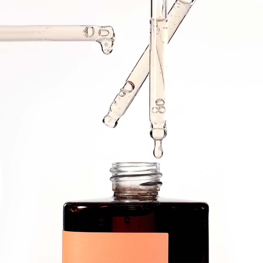 Close-up of multiple serum droppers dripping the light amber liquid of the Missha Time Revolution Primestem 100 Lifting Serum into the bottle's neck. This highlights the concentrated texture of the anti-aging serum for firming.